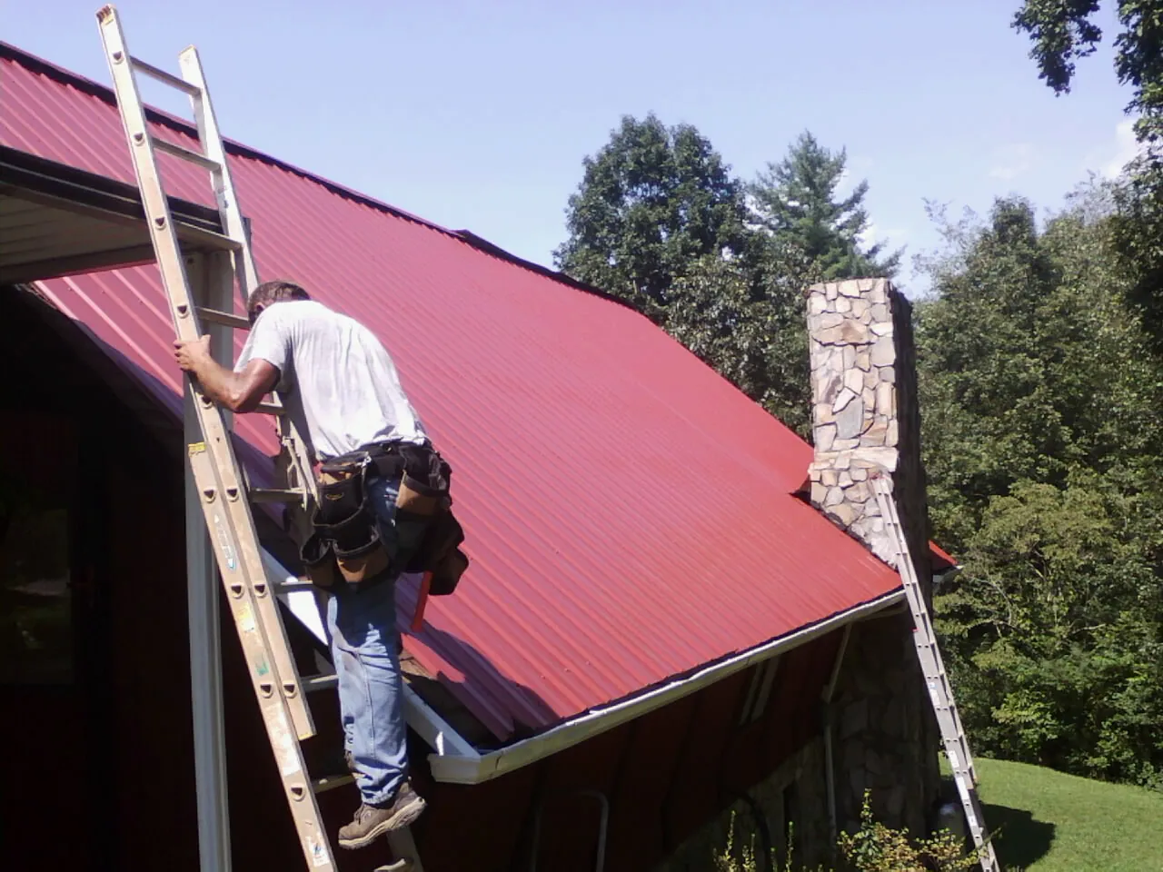 Roofer on a ladder beside a red metal roof and stone chimney — Soffit & Fascia Repair by Roofing Brothers in Lascassas